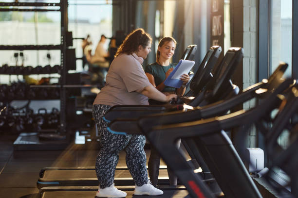 A personal trainer assisting a woman on a treadmill in a gym, discussing a structured weekly workout plan for steady progress.