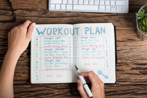 A top view of a person's hands holding a pen, creating a colorful workout plan in a notebook, placed on a wooden desk with a keyboard and a small plant nearby.