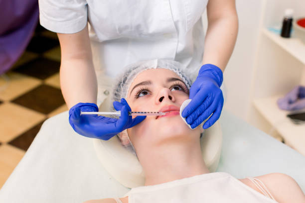 A close-up of a woman lying down, wearing a protective cap, while a practitioner in gloves injects a cosmetic filler or treatment into her lips in a clinic setting.