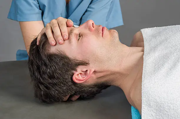 A man lying on a table with a professional providing care by supporting his forehead during recovery.