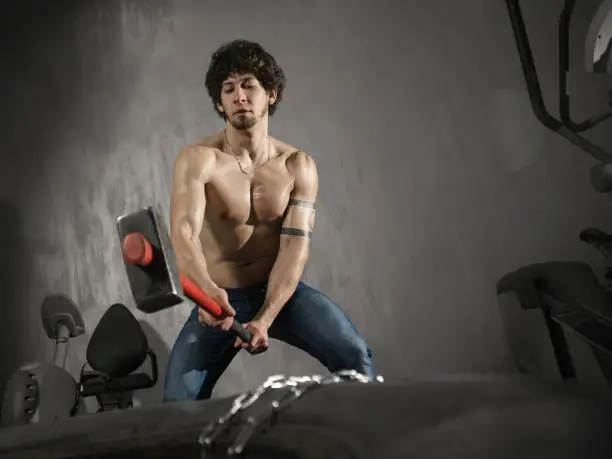 A shirtless man striking a large tire with a red-handled sledgehammer in a modern gym environment.