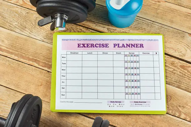 An exercise planner on a wooden surface surrounded by dumbbells and a water bottle.