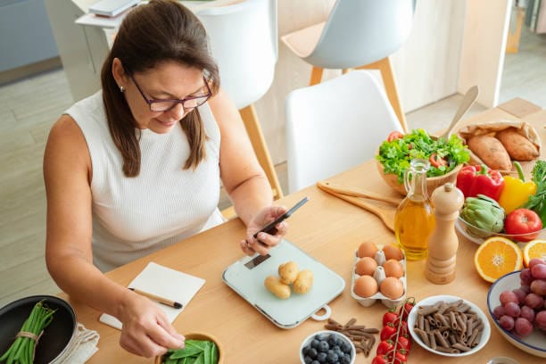 Woman measuring and preparing healthy ingredients at a table filled with fruits, vegetables, and other nutritious foods.