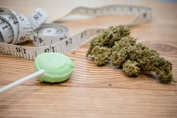 A close-up image of cannabis buds, a green cannabis-infused lollipop, and a measuring tape on a wooden surface, symbolizing the relationship between weed consumption, calories, and weight management.