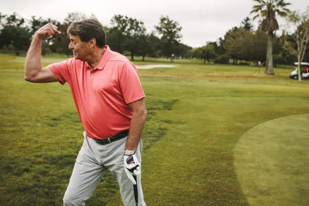 Senior golfer in a pink shirt flexing his arm on the golf course, showcasing the benefits of strength training for better performance.